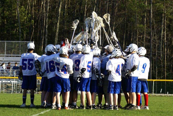 A lacrosse team in white and blue uniforms huddling and raising their sticks on a green field with trees in the background